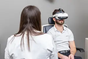 A man is sitting on a chair with a virtual reality headset, and a woman is standing behind him in a room. The man is wearing a white polo shirt and a black belt, and the woman is wearing a white coat. The man is looking at the woman, and the woman is looking at the man. Behind them is a gray wall, and a white object is on the right side of the wall.
