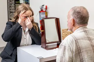 A woman is putting in a contact lens while an older man watches her in a room with a fire extinguisher and mirror on a desk.