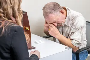 An elderly man wearing a plaid shirt is sitting at a table with a woman wearing a black shirt. He is resting his head on his hand, which is covering his face. He is looking at the woman who is probably looking at something. The table has a wooden box with a lock and some white bottles. Behind them is a white wall.