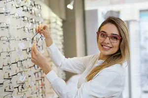 A smiling woman in a white shirt holding a pair of glasses while looking at a wall of glasses in a shop