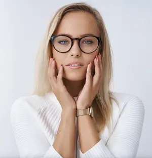 A blonde woman wearing glasses and a white sweater with her hands on her cheeks