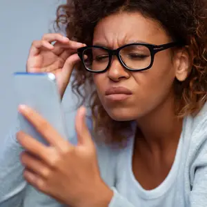 A woman with curly hair squinting at her phone and adjusting her glasses
