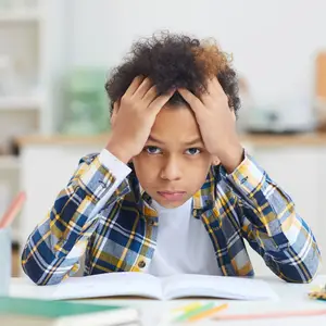 A young boy with curly hair, wearing a plaid shirt, is sitting at a desk with his hands on his head, looking at a book and seems to be studying.