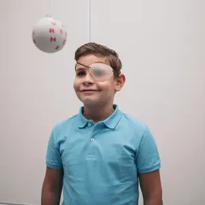 A smiling boy wearing a blue shirt and an eye patch is standing in a white room and looking at a ball with red letters on it.