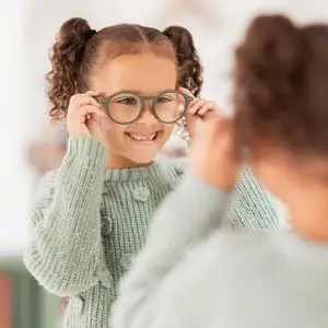 A little girl is trying on a pair of glasses in front of a mirror.