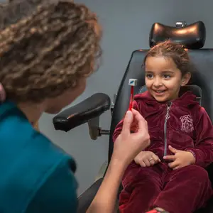 A smiling child sits in a dental chair while a dental professional brushes their teeth