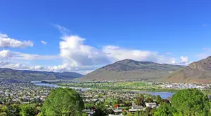 A view of a town with a river and mountains in the distance under a blue sky with white clouds.