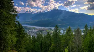 A view from the top of a hill shows a mountain range, a river, and a town surrounded by trees and a blue sky with clouds.