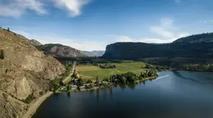 Aerial view of a river flowing through a valley surrounded by mountains, with a green field and houses near the water.
