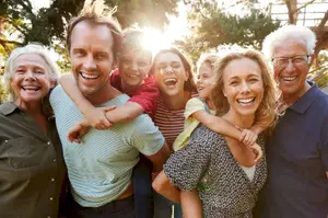 A group of people, including a woman, a man, and children, are smiling and posing for a photo in a garden with trees. The man is carrying a child on his shoulders, and the woman is holding a child's hand. The man in the middle is looking at the camera, and the children are looking at something. Behind them is a ladder leaning against a tree, and there is a house with a roof. 
