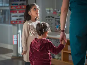 A woman in a teal blue scrub suit holds the hand of a young girl in a red hooded sweatshirt, while another girl in a white sweater with sequins on the collar stands behind them in a room with shelves of sunglasses and other items in the background.