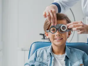A young boy is being checked for his vision by a doctor in a blue chair wearing a denim jacket