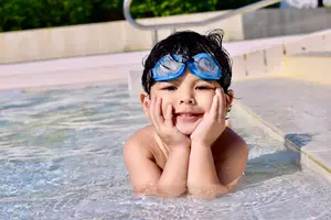 A young boy wearing goggles and a yellow shirt is smiling in a swimming pool with blue water and a concrete deck.