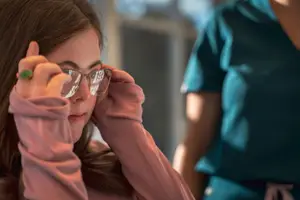 A woman wearing glasses and a ring is adjusting her glasses with her left hand while looking at something in front of her. Behind her, a person wearing a blue shirt and pants is standing and looking at her. The background is blurry.