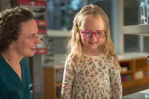 A young girl with glasses and a lady in green scrubs are smiling at each other in a room