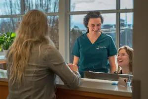 Three women stand behind a counter and speak to one another. One woman is wearing glasses and a green shirt, and the other two are wearing glasses as well.
