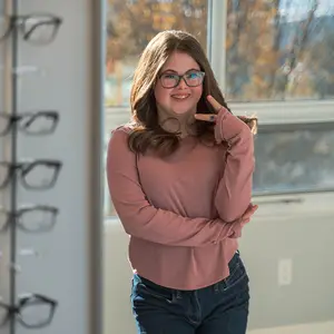 A woman is standing in front of a display of eyeglasses, smiling and posing for a picture with her hand on her forehead. She is wearing a pink long-sleeve shirt and blue jeans. Behind her, there is a glass window with a view of trees and a building. A white wall with a rack of eyeglasses is on the left side. A white power outlet is mounted on the wall on the right side.