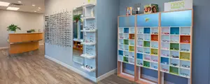 Interior of a modern optometry store with blue walls, wooden floors, and displays of glasses, a potted plant, and a reception desk. The shelves hold an array of glasses in various colors and styles, and the walls are adorned with mirrors and framed pictures.