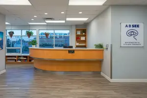 An empty reception desk inside an optometrist's office with a glass window and a wooden cabinet, with a signboard on the wall.