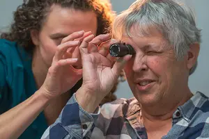 A woman wearing glasses looks through an otoscope, a device used to examine the ear. A medical professional, dressed in a blue shirt, holds the otoscope in place and examines the woman's ear. The woman has her eyes closed and appears focused on the examination.