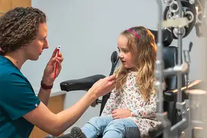 A girl sits in a chair while an eye doctor looks at her eyes with a light.