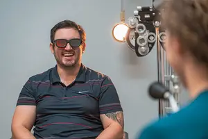 Man in a black shirt with red stripes, wearing black glasses, and smiling at a person wearing a blue shirt in front of an eye exam machine.