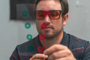A man wearing orange goggles adjusts a red object in a blurred background with green circles.