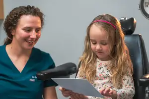 A girl in a dental chair holds a tablet while a woman in a blue lab coat smiles beside her.