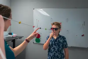 A woman holding a stick with a yellow ball at the end is explaining something to a boy wearing glasses and a blue floral shirt in front of a whiteboard.