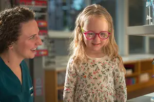 A smiling young girl with pink glasses stands in a hospital room, looking at a woman in a green scrubs who is smiling back at her.