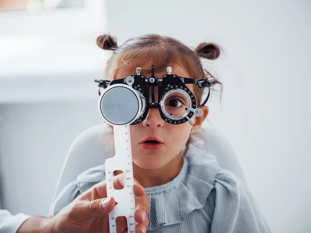 Young girl wearing glasses and looking into a device that measures her vision, held by an adult in a white coat.