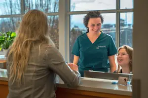 A woman standing behind a reception counter with a smiling woman in front of her and another woman standing beside her wearing glasses and smiling with a potted plant behind her