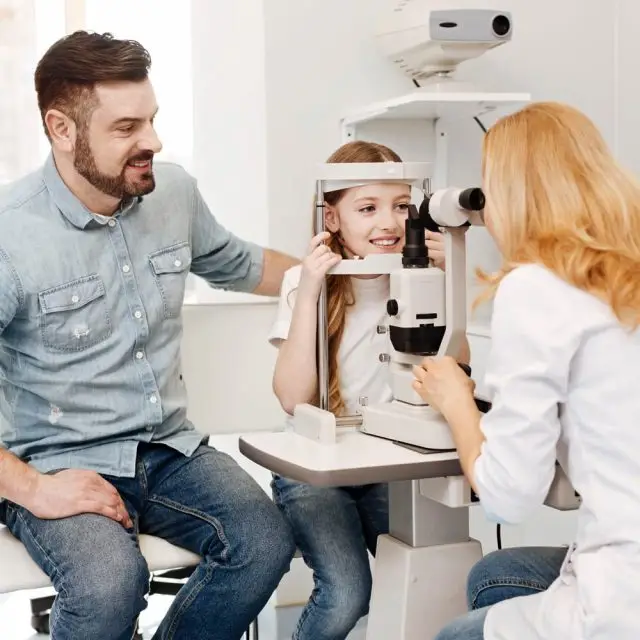 A young girl is having her eyes checked by a doctor and her father is watching.