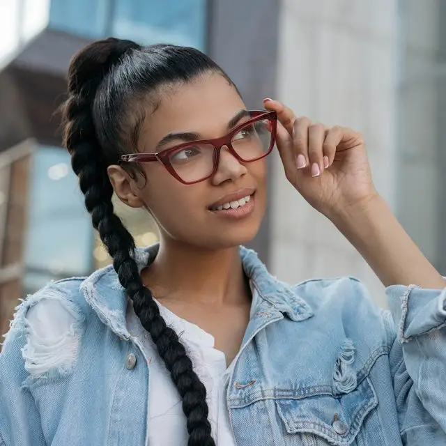 A woman with a braided hairstyle wearing red-framed glasses and a denim jacket, standing in front of a building.