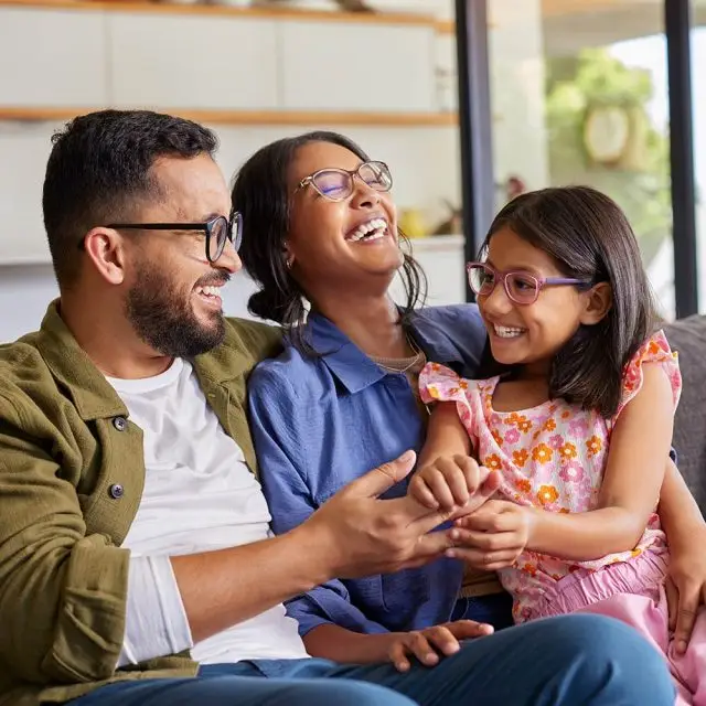 A man, a woman, and a young girl wearing glasses are sitting together on a couch, laughing and smiling, with a window and plants visible in the background.