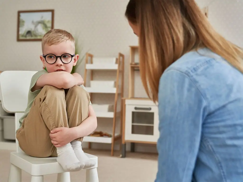 A young boy wearing glasses and a green shirt is sitting on a chair with his arms crossed, while a woman in a blue shirt looks at him. Behind them is a wooden cabinet with a picture frame on the wall, a wooden shelf, and a white chair. The room has a white wall and a carpeted floor.