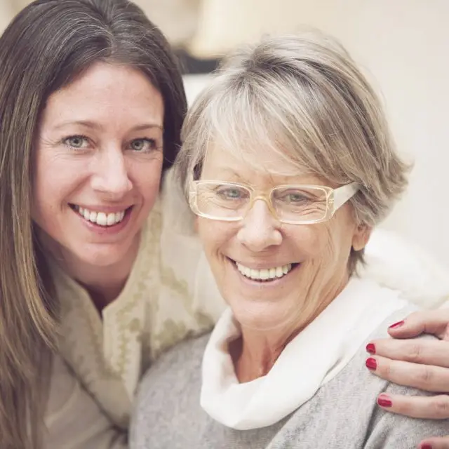 Two smiling women posing for a photo, one with glasses and a white scarf, the other with red nail polish.