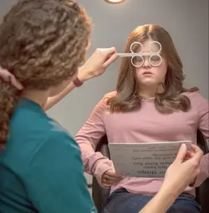 A woman in a pink long-sleeve shirt and jeans is having her eyes checked by an optician in a clinic. The woman is sitting in a chair while the optician is holding a paper and a pen. The optician is wearing a green shirt and a watch. The room is dimly lit, and a lamp is on the ceiling. The woman is holding a pair of scissors and looking at the optician.