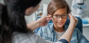 A young boy is adjusting his eyeglasses with the help of an adult in a clinic setting