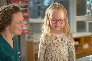 A woman in green scrubs speaks with a young girl wearing pink glasses in a hospital setting