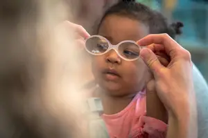 A young girl in a pink shirt tries on a pair of glasses while sitting in a chair.