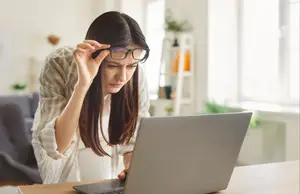 A woman wearing glasses is looking at her laptop and holding her head in her hand