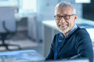 An older man wearing glasses, a blue ID, and a blue sweater is smiling while sitting on a chair in an office with a computer and a desk behind him.