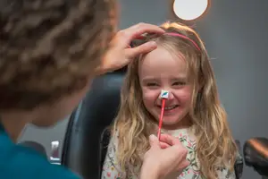 A young girl smiles and holds a toothbrush in her mouth as a woman adjusts her hair behind her head, sitting in a dental chair with a light overhead.
