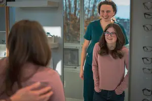 Three women in a room, one of them is a smiling woman standing in front of a mirror with two other women behind her smiling at her in an optical shop.