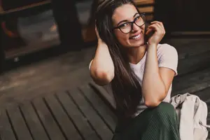 A woman sitting on a wooden floor wearing glasses and a backpack smiling for a photo