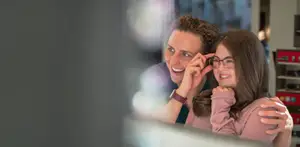 A man and a woman are smiling and looking into a mirror with a reflection of another person in the mirror.