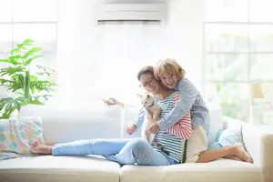A family of two women and a child with a dog sitting on a white couch in a living room with an air conditioner, plant, and lamp.
