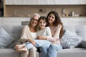 A family of three, an elderly woman, a young woman, and a young girl, sitting on a couch and smiling while looking at the camera.