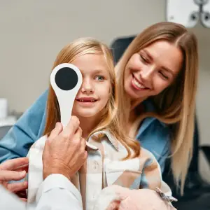 Girl getting her eye checked by an optometrist with her mother smiling in the background
